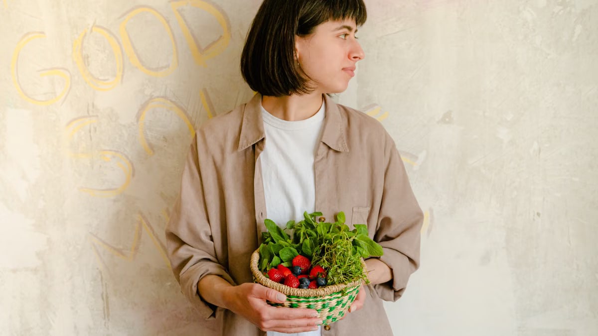 Woman standing with a basket of fresh greens and berries against a rustic wall background.
